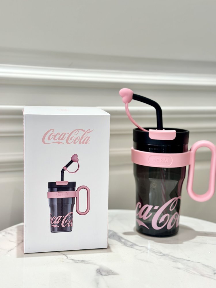 Black Coca-Cola tumbler with pink handle and straw next to a white box with Coca-Cola branding.