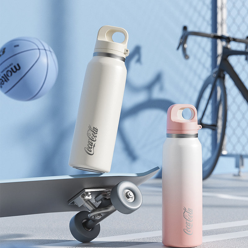 Two Coca-Cola water bottles, one white and one pink, on a skateboard with a basketball and bicycle in the background.
