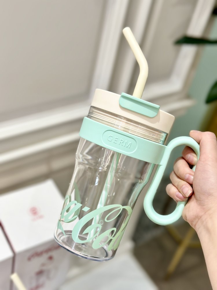 Clear tumbler with mint green lid and handle, held by a hand indoors.