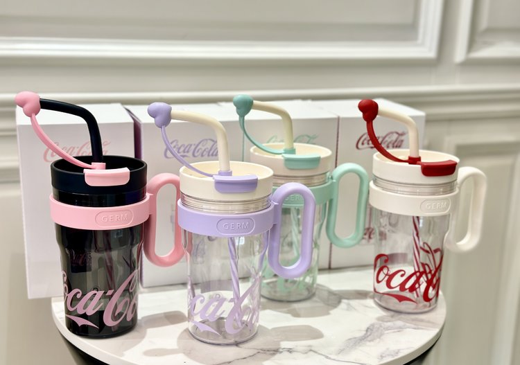 Set of Coca-Cola branded cups with handles and straws on a white surface.