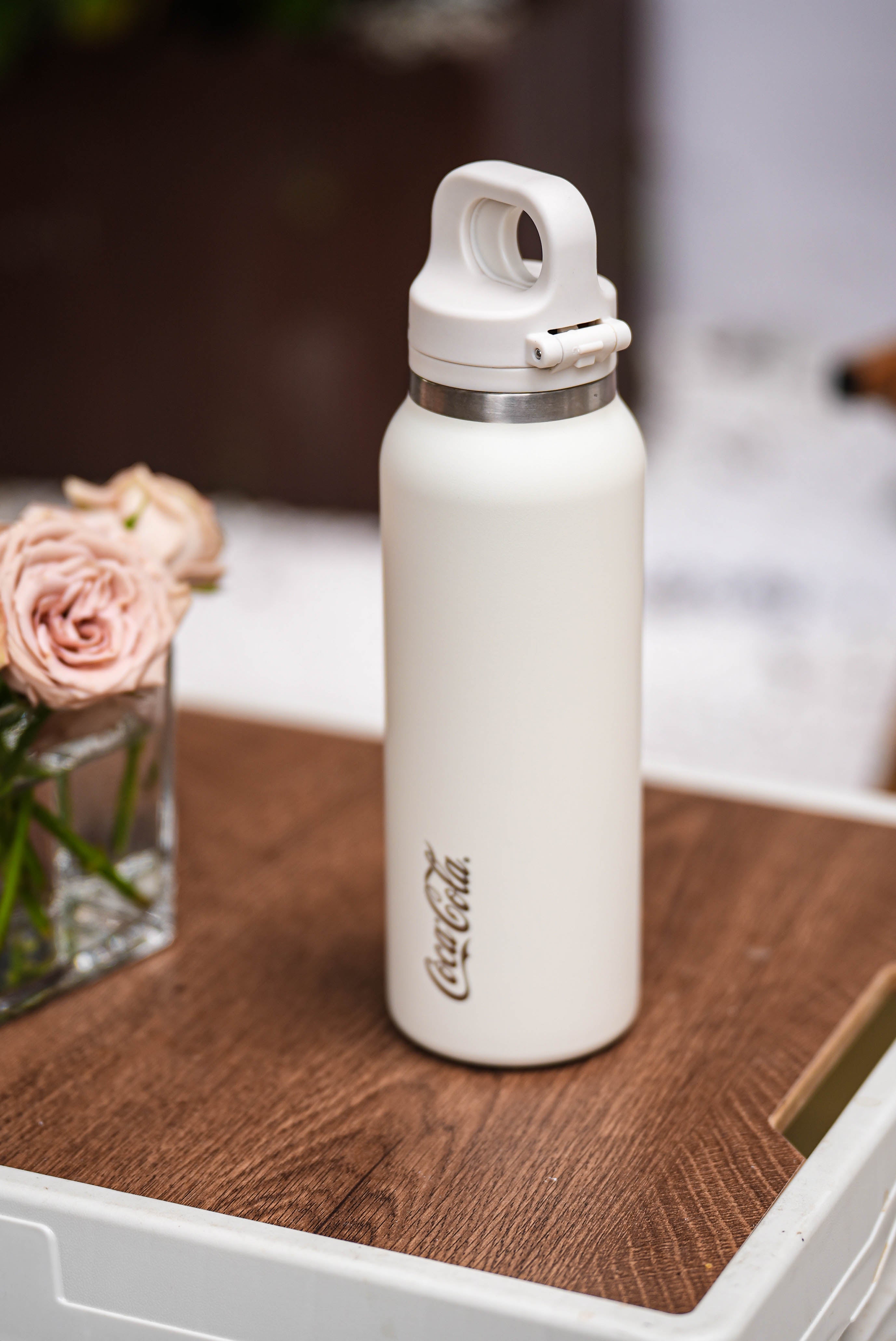 White Coca-Cola water bottle on a wooden surface with pink flowers in the background