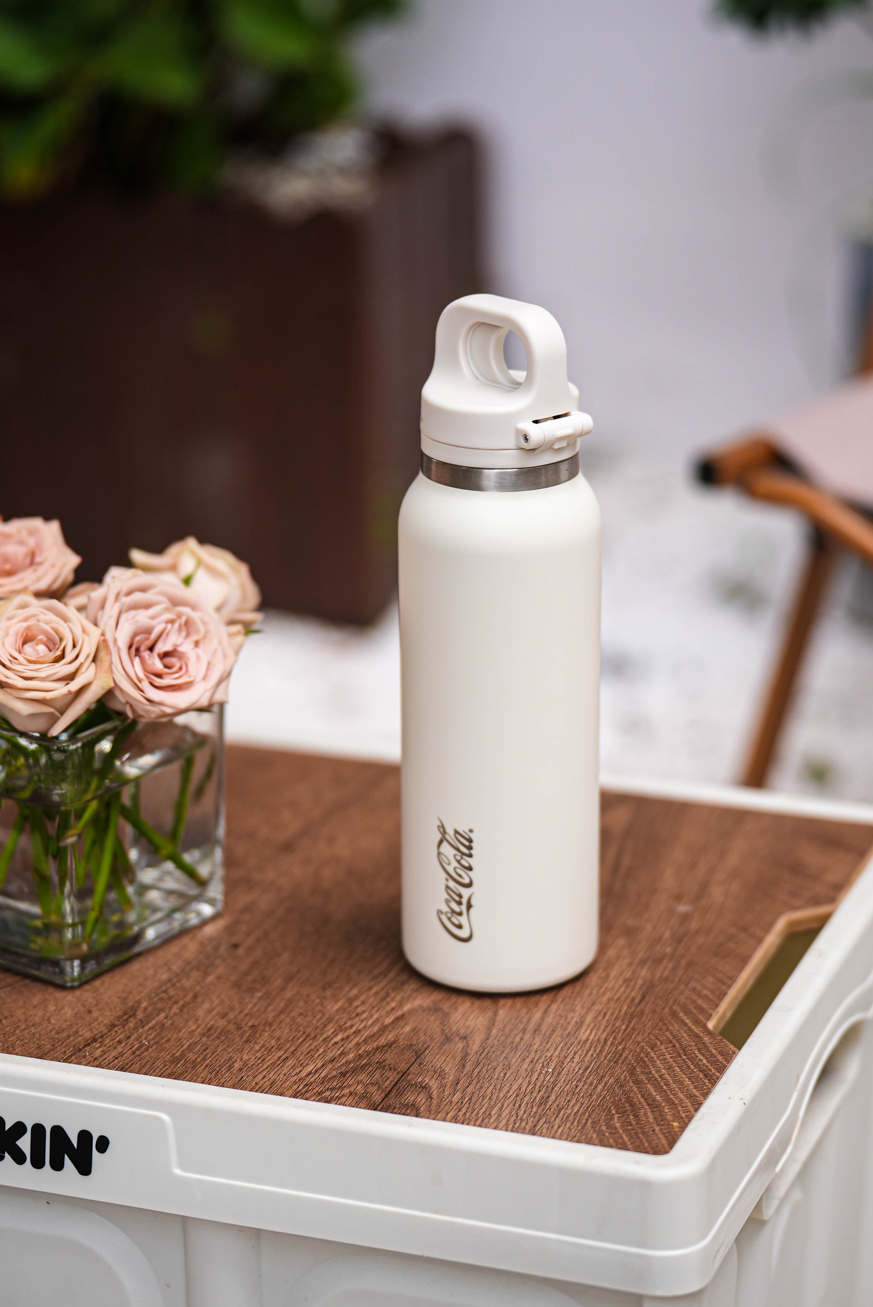 White water bottle with Coca-Cola logo on a wooden surface next to a vase of flowers.