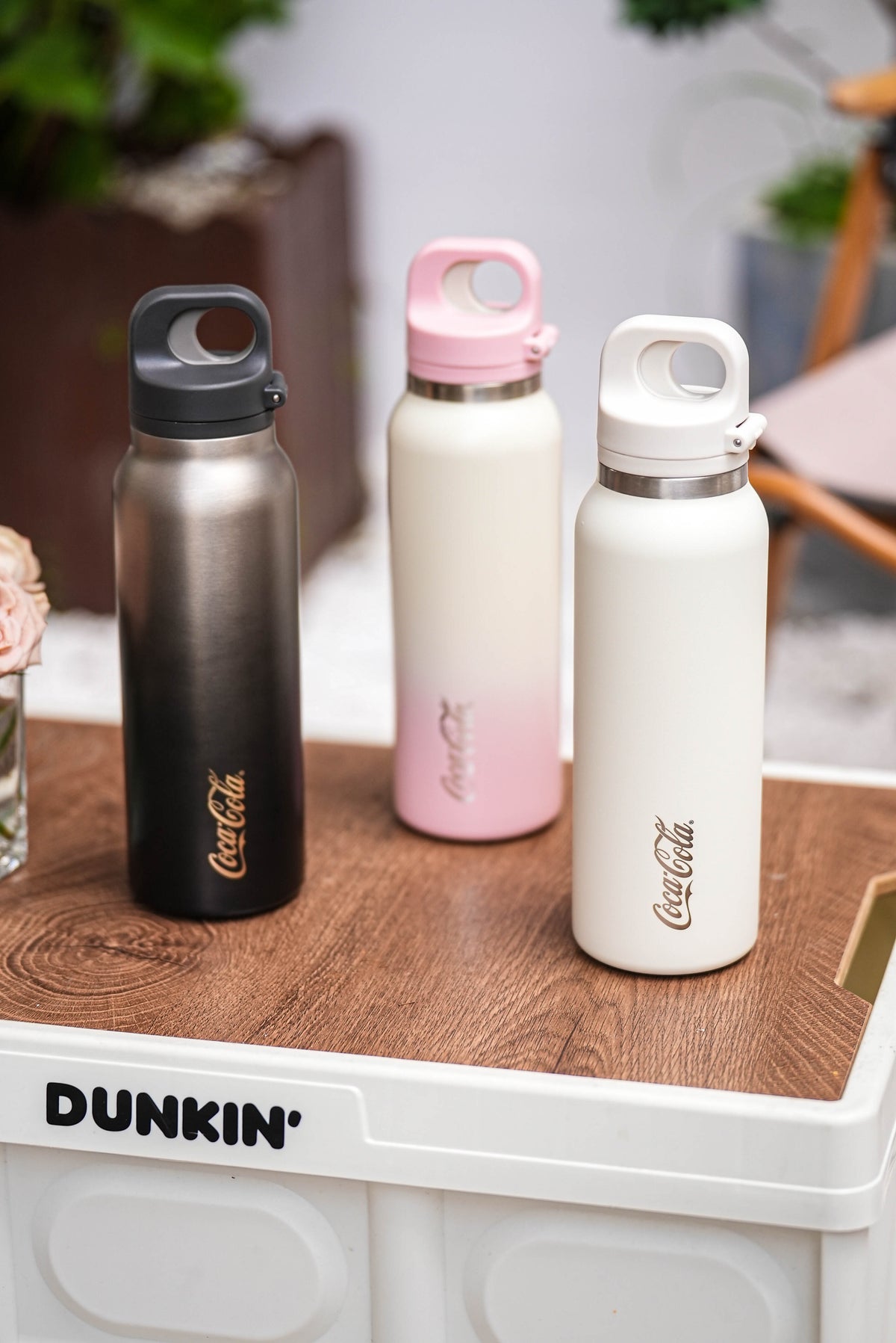 Three Coca-Cola water bottles on a wooden surface with a Dunkin' sign in the background.